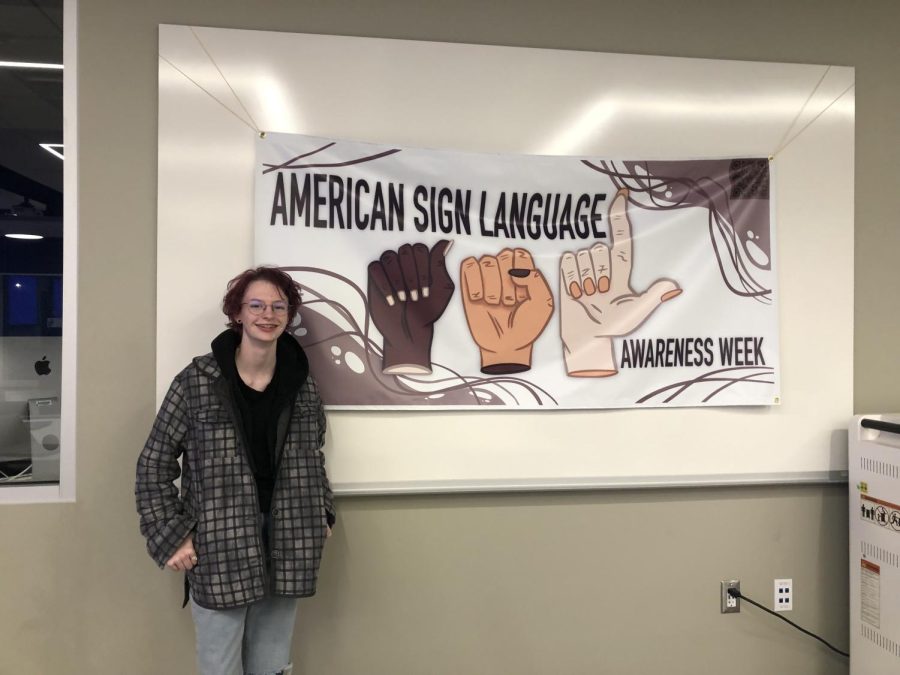Em Burch posing next to the banner they designed for a week of ASL awareness.